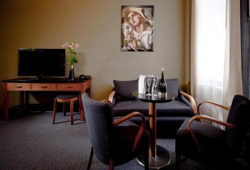 A cozy hotel room features a comfortable seating area with a small table set for two, a desk with a TV, and a serene painting on the wall, ideal for travelers seeking relaxation and a touch of elegance during their stay.
