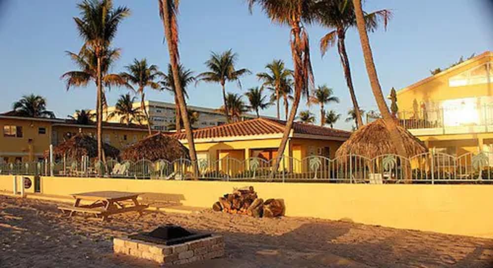 A serene beachside resort features palm trees, thatched huts, and picnic tables under a clear sky, ideal for relaxing tropical vacations.