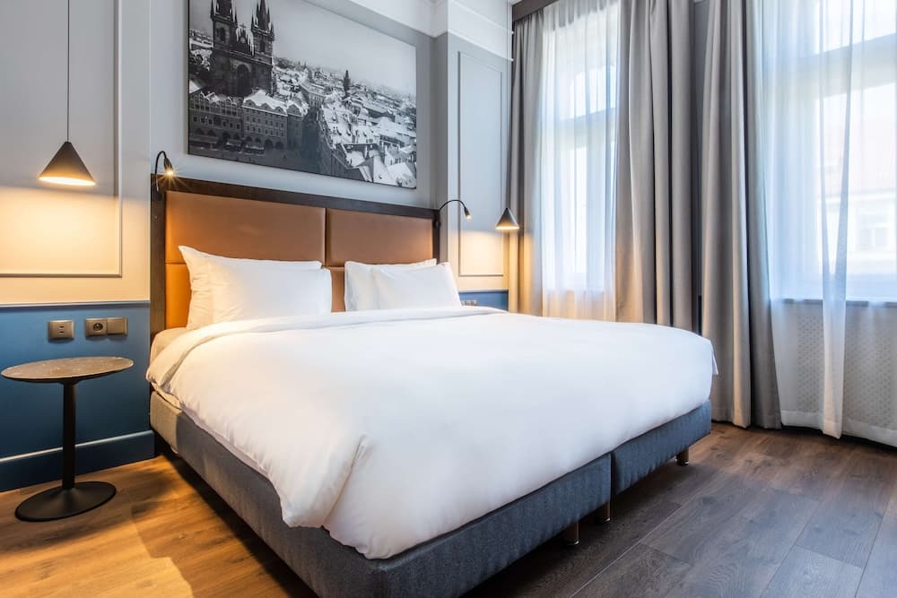A cozy hotel room features a neatly made bed with white linens, a stylish leather headboard, soft lighting, and a cityscape artwork, creating an inviting atmosphere ideal for travelers seeking comfort and relaxation.