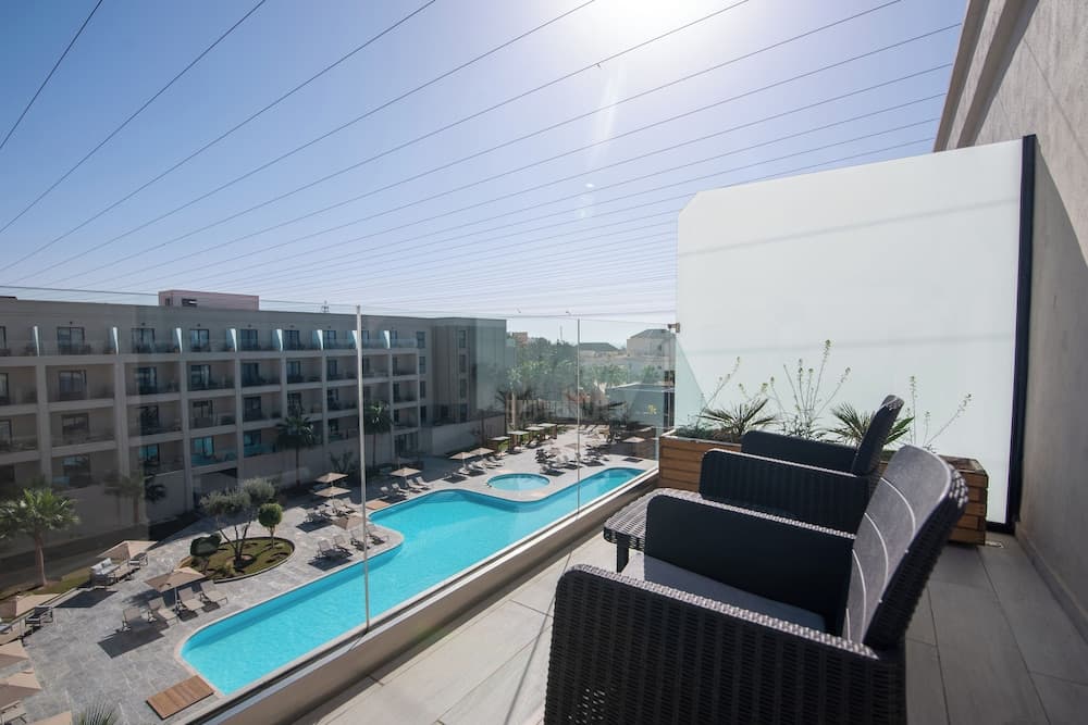 A serene hotel balcony overlooks a vibrant blue pool surrounded by lounge chairs and palm trees under a clear sky, ideal for travelers seeking relaxation and leisure.