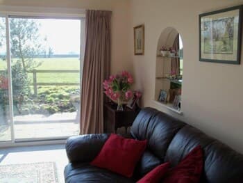 A cozy living room with a leather sofa adorned by red cushions overlooks a serene garden through a glass door, ideal for travelers seeking a tranquil countryside retreat.