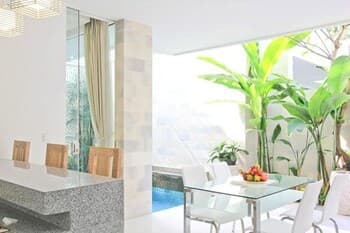 A serene indoor-outdoor dining area bathed in natural light features modern furniture, lush greenery, and a tranquil pool view, ideal for a relaxing vacation retreat.