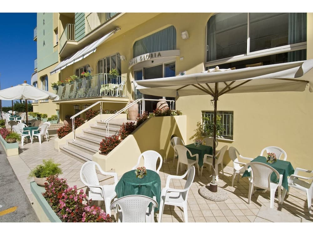 A sunny outdoor terrace at Hotel Gloria features inviting white chairs, green tables, and large umbrellas, creating a relaxed and welcoming atmosphere ideal for travelers seeking leisure and comfort.