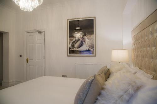 A cozy hotel room features a plush bed adorned with soft pillows and a fur throw, complemented by elegant decor including a chandelier-framed artwork and warm lighting, ideal for travelers seeking comfort and luxury.