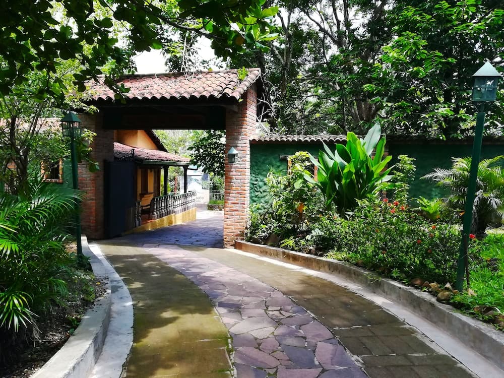 A serene tropical garden pathway lined with lush greenery and vibrant plants leads to a charming brick archway entrance, ideal for travelers seeking a tranquil retreat in a natural setting.