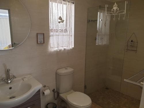 A cozy bathroom with beige tiles, a glass shower enclosure, a white toilet, and a sink under a circular mirror, featuring lace curtains and a butterfly decoration, ideal for travelers seeking a comfortable and private accommodation space.