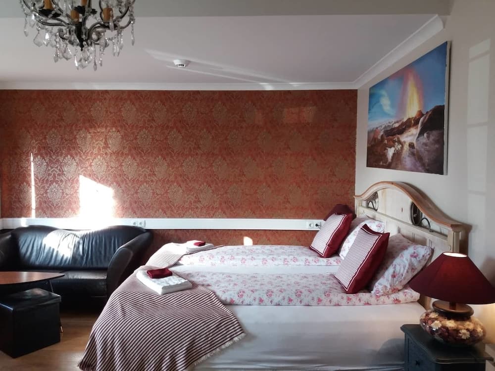 A cozy hotel room features a floral-patterned bed, elegant chandelier, and vibrant red wallpaper, creating a warm and inviting atmosphere ideal for travelers seeking comfort and charm.