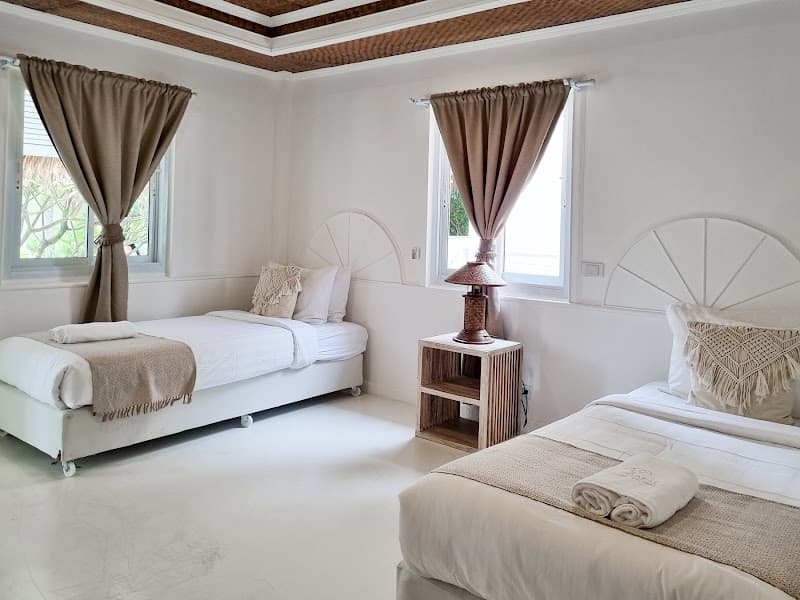 A serene and cozy bedroom at Elephant Beach Club & Resort Samui features two neatly made beds with white linens, a wooden bedside table with a lamp, and large windows draped with beige curtains, ideal for travelers seeking relaxation and comfort near Chaweng Beach.