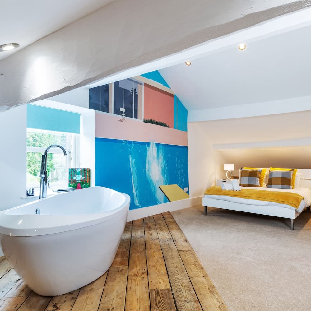 A cozy attic bedroom with a freestanding bathtub, vibrant blue wall mural, wooden flooring, and a neatly made bed creates a serene and stylish retreat ideal for travelers seeking unique accommodations.