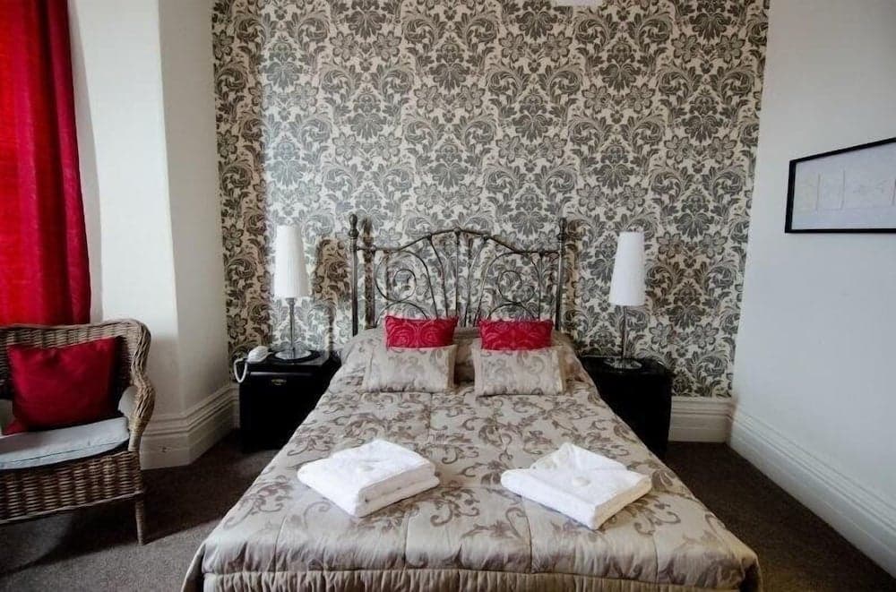 A cozy hotel room features a bed with ornate bedding and red accents against a patterned wallpaper backdrop, complemented by a wicker chair and warm lighting, ideal for travelers seeking comfort and charm.