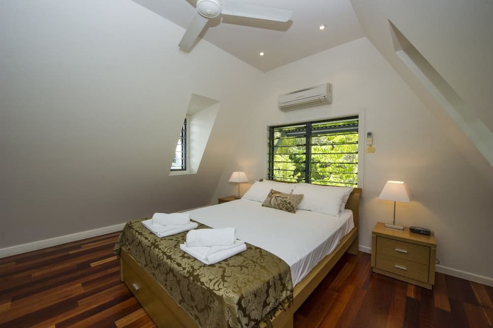 A cozy attic bedroom with sloped ceilings, wooden flooring, a neatly made bed adorned with folded towels, bedside lamps, and a window offering a serene view of greenery, ideal for travelers seeking a tranquil retreat.