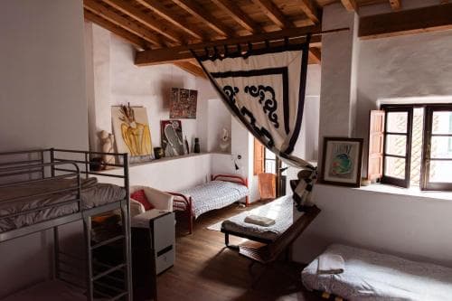 A cozy hostel room with wooden beams, bunk beds, and eclectic decor bathed in warm sunlight through open shutters, ideal for budget travelers seeking a charming and artistic atmosphere.