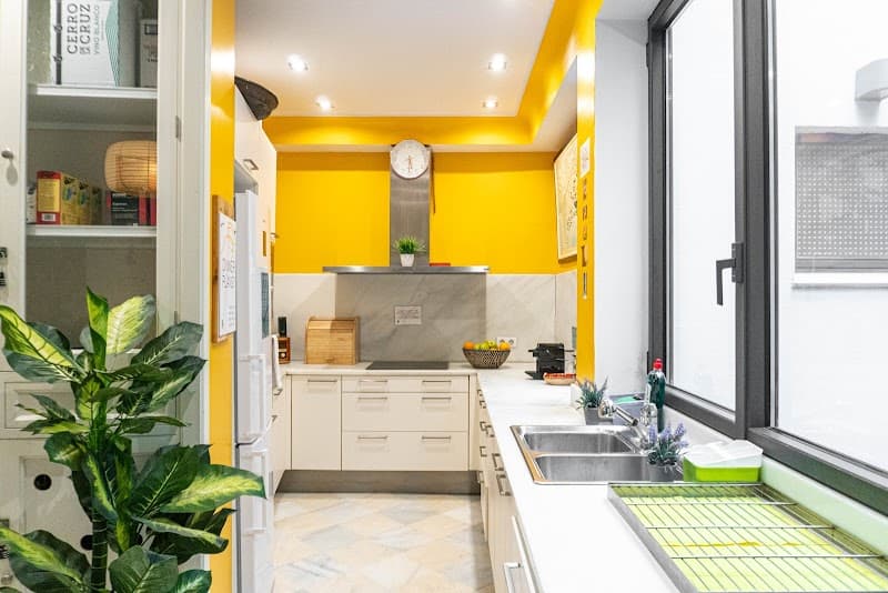 A vibrant yellow-walled kitchen in La Banda Rooftop Hostel in Sevilla, Spain, exudes warmth and modern charm with sleek white cabinets, a granite countertop, and natural light streaming through large windows, ideal for travelers seeking a cozy and stylish cooking space.