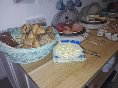 A cozy breakfast buffet scene featuring a wooden table laden with fresh bread rolls in a woven basket, assorted cheeses, meats under glass domes, and neatly arranged utensils, perfect for a relaxing morning getaway or hotel stay.