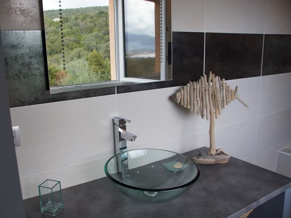 A serene modern bathroom features a glass vessel sink, a driftwood fish sculpture, and a large window overlooking lush greenery, ideal for eco-friendly retreats.