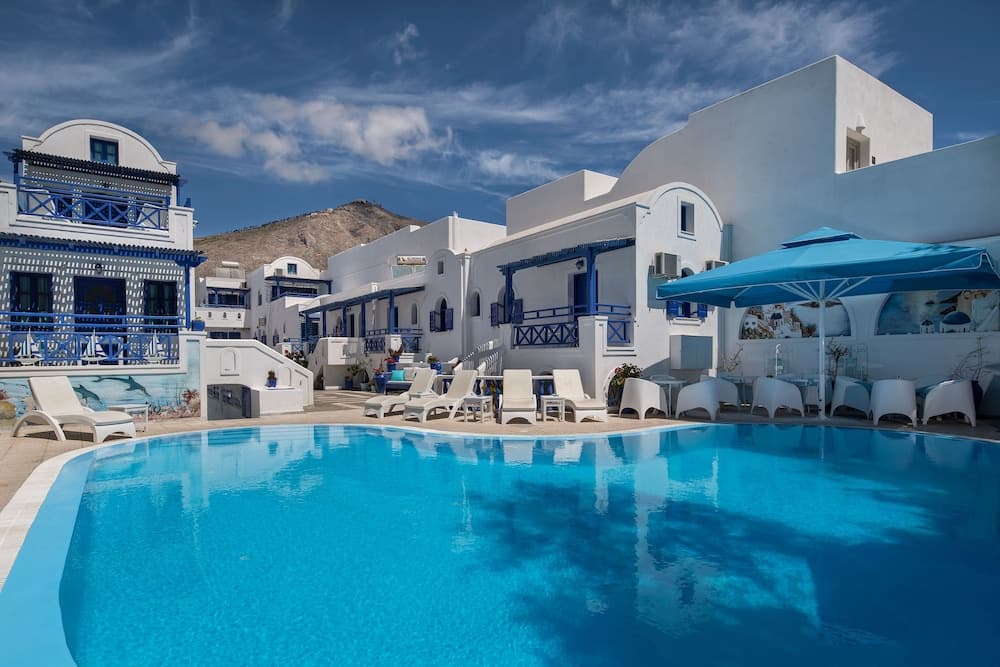 A serene Mediterranean resort features a sparkling blue pool surrounded by white sun loungers, under a clear sky with traditional white buildings and blue accents, ideal for luxury vacation seekers.