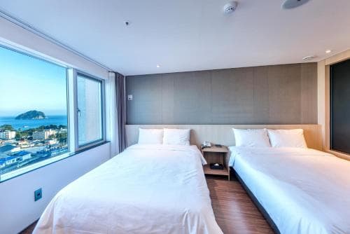 A cozy hotel room with two neatly made beds overlooks a scenic coastal view through a large window, ideal for travelers seeking relaxation and comfort by the sea.