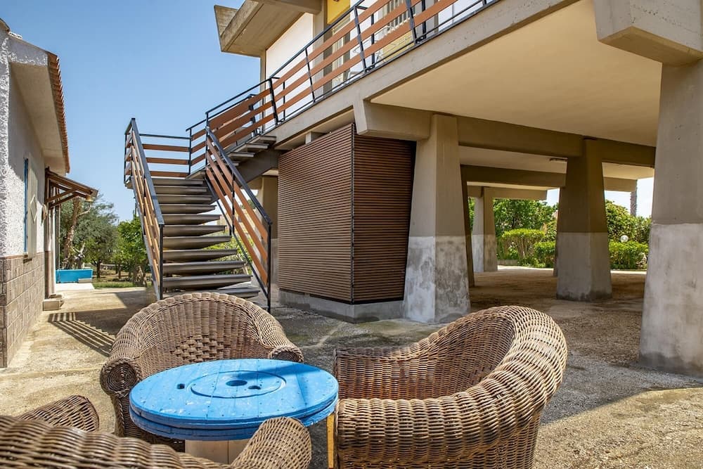A serene outdoor patio features wicker chairs around a blue table under a modern staircase, ideal for relaxing in a tranquil residential setting.