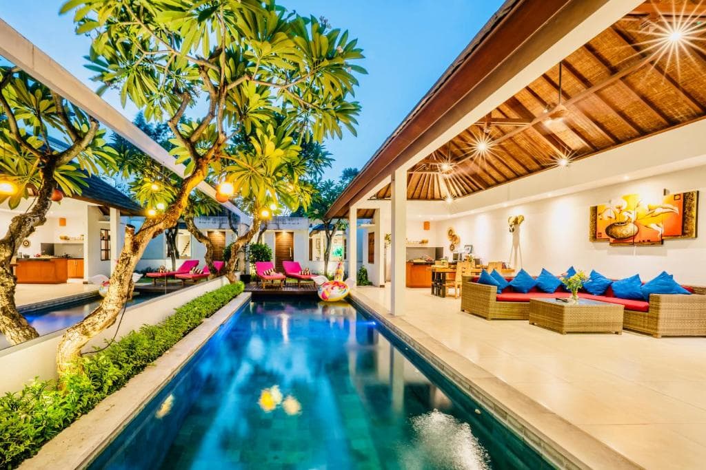 A serene tropical villa features a tranquil pool lined with lush greenery and string lights, complemented by cozy lounge areas under a wooden-beamed ceiling, ideal for a luxurious vacation retreat.