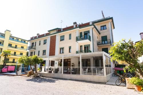A charming multi-story apartment building with balconies and a covered patio area surrounded by greenery offers a cozy retreat in a sunny urban setting, ideal for travelers seeking comfortable accommodations near local attractions.