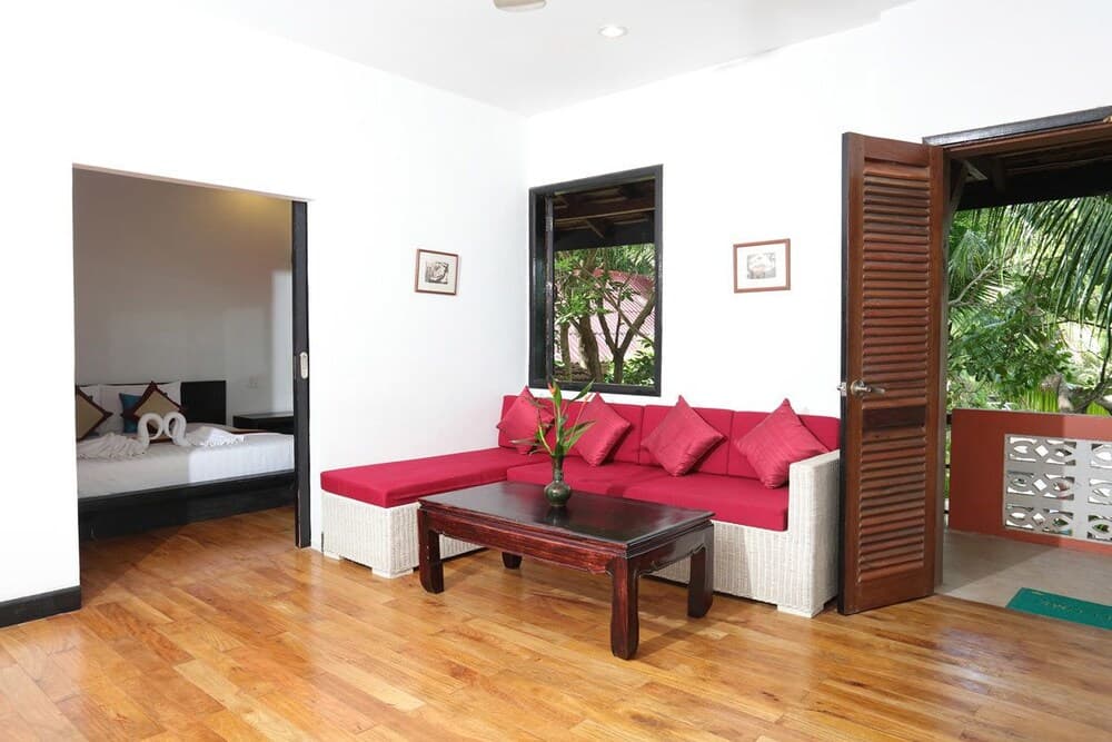 A cozy tropical villa living area features a vibrant red sofa set against white walls, wooden flooring, and an open door leading to lush greenery, ideal for travelers seeking a serene and stylish retreat.