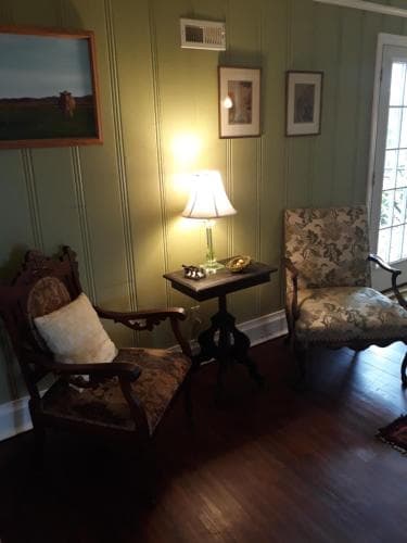 A cozy vintage living room with green-paneled walls, antique furniture, and soft lighting creates an inviting atmosphere perfect for a relaxing getaway or heritage tourism experience.