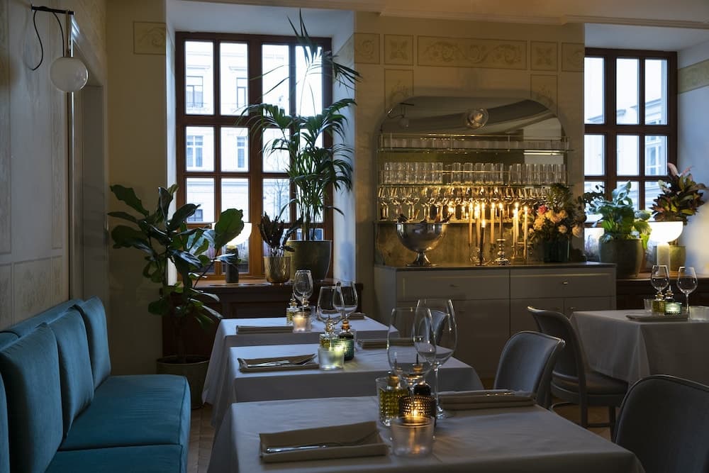 A cozy, elegantly set restaurant interior features white tablecloths, candlelight, lush green plants, and large windows, creating an intimate dining atmosphere perfect for romantic evenings or special occasions.