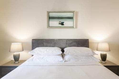 A serene hotel room features a neatly made bed with white linens, flanked by bedside lamps and a tranquil landscape painting above the headboard, ideal for travelers seeking relaxation and comfort.