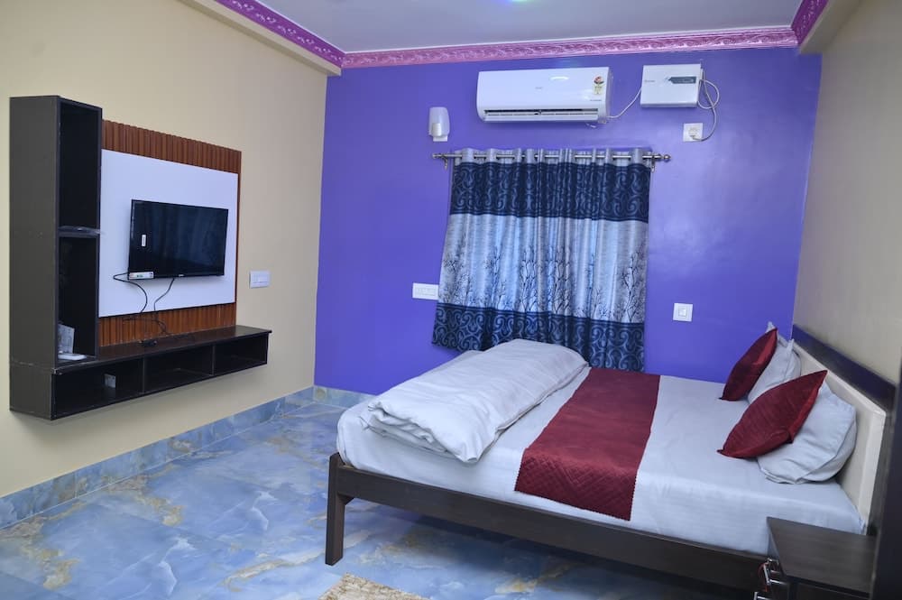 A cozy hotel room features a neatly made bed with red accents, a purple wall, patterned curtains, a mounted TV, and marble flooring, ideal for travelers seeking comfort and relaxation.