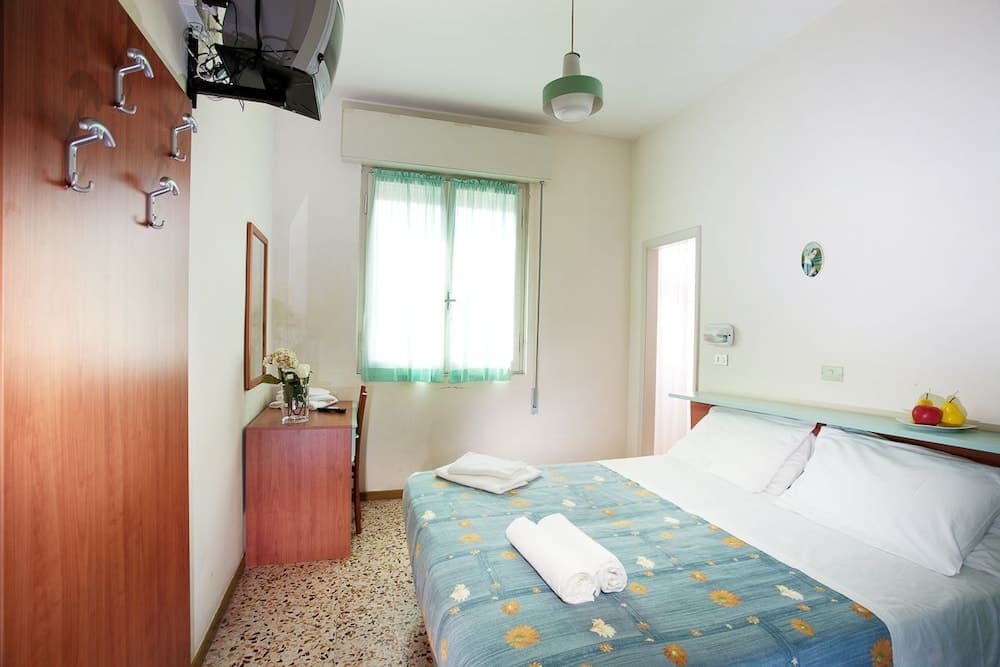 A cozy hotel room features a neatly made bed with a floral blanket, soft lighting from a green pendant lamp, and a window with sheer curtains, ideal for travelers seeking comfort and simplicity.