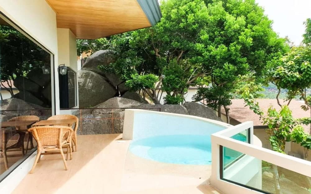 A serene outdoor patio features a tranquil pool surrounded by lush greenery and large rocks, ideal for relaxing getaways with wicker furniture and a cozy wooden overhang.