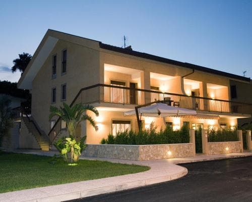 A cozy evening view of a warmly lit two-story villa with balconies, surrounded by lush greenery and palm trees, evokes a serene vacation atmosphere ideal for travelers seeking relaxation.
