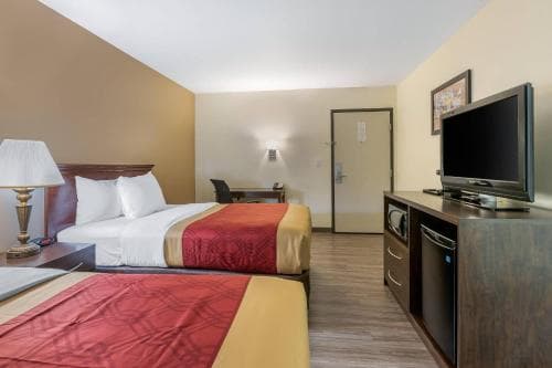 A cozy hotel room features two neatly made beds with red and yellow bedding, a flat-screen TV on a wooden dresser, and a small desk area, ideal for travelers seeking comfort and convenience during their stay.