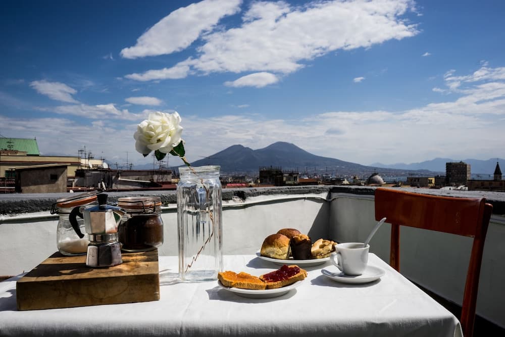 A serene rooftop breakfast setup with a white rose, coffee maker, pastries, and a stunning view of distant mountains under a partly cloudy sky, ideal for travelers seeking a peaceful morning retreat.