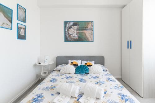 A cozy bedroom with a neatly made bed adorned in floral bedding, blue accents, and beach-themed artwork creates a serene vacation atmosphere ideal for travelers seeking relaxation.