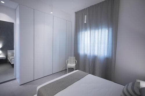 A serene minimalist bedroom features a neatly made bed, sleek white wardrobes, and soft gray curtains, creating a tranquil atmosphere ideal for travelers seeking a peaceful retreat.