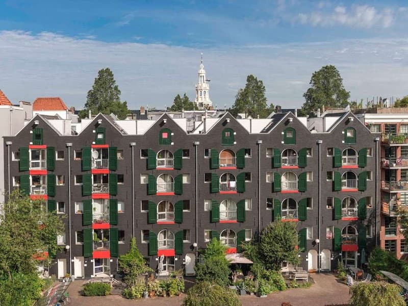A vibrant urban scene featuring the Ibis Amsterdam Centre Stopera hotel with its distinctive black facade, green shutters, and red accents, set against a backdrop of lush greenery and a historic church spire, ideal for travelers seeking a charming stay in the heart of Amsterdam.