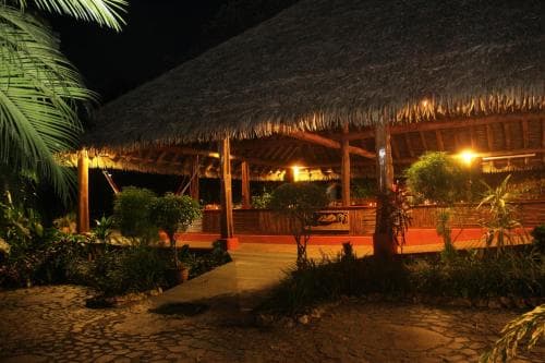 A cozy, thatched-roof bungalow illuminated by warm lights at night, surrounded by lush tropical plants, creates an inviting atmosphere ideal for a serene vacation getaway.