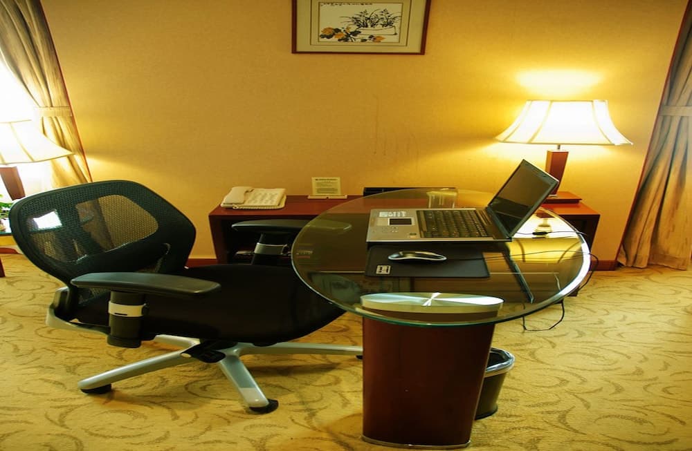 A cozy hotel room workspace features a glass-top table with a laptop, ergonomic chair, warm lighting from a desk lamp, and a serene ambiance ideal for business travelers seeking productivity.