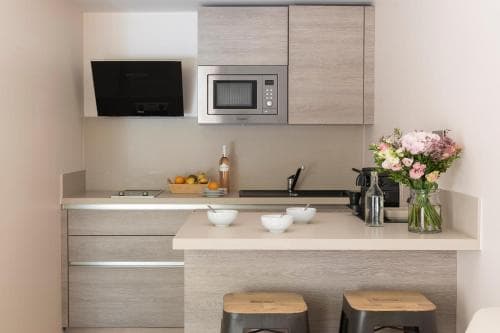 A cozy modern kitchenette with sleek wooden cabinets, a microwave, and a vase of fresh flowers creates an inviting atmosphere perfect for travelers seeking comfort in a compact apartment setting.