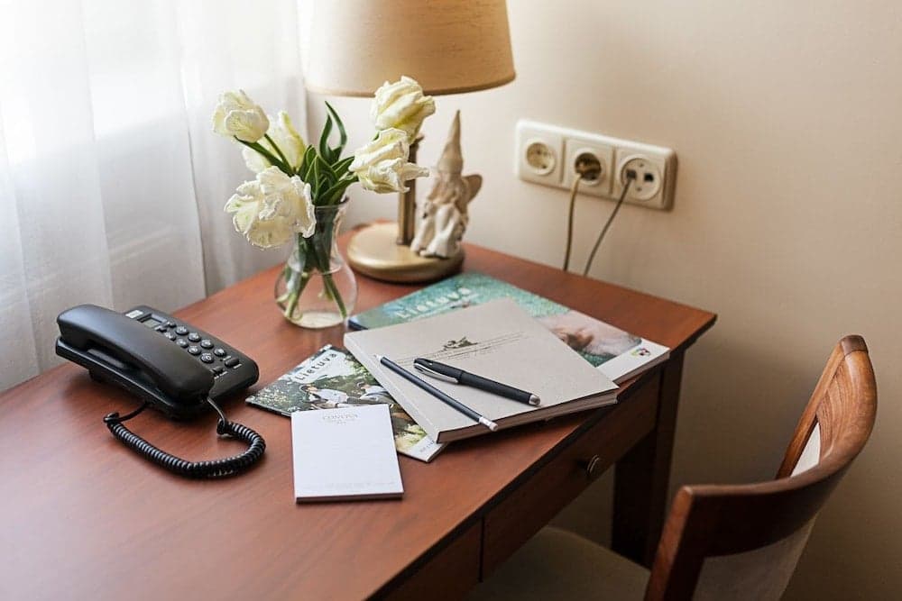 A cozy hotel room desk setup features a wooden table adorned with white tulips, books, stationery, and a telephone, creating an inviting atmosphere ideal for travelers seeking a comfortable workspace.