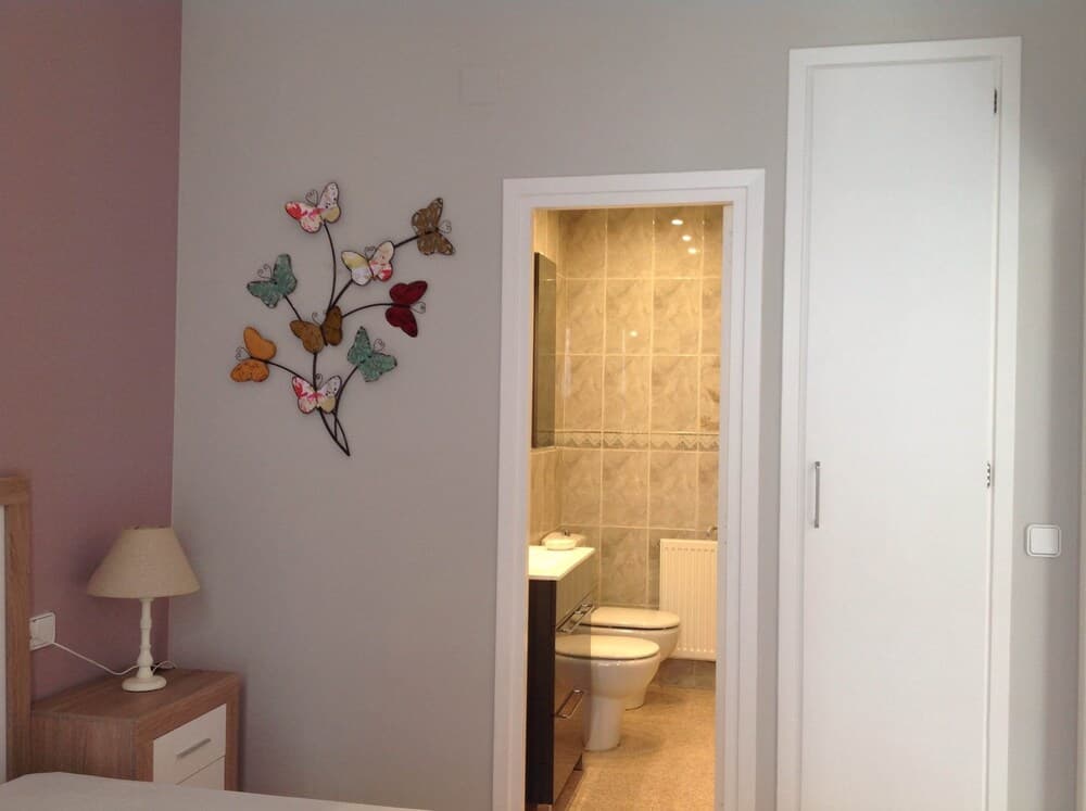 A cozy bedroom corner with a butterfly wall art and bedside lamp leads to an inviting bathroom with beige tiles, perfect for travelers seeking comfort and style in their accommodations.