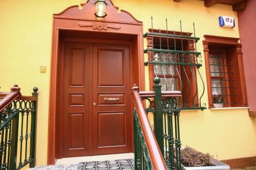 A charming yellow building entrance with a wooden door, ornate green railings, and decorative window grills evokes a cozy, inviting atmosphere perfect for travelers seeking a quaint accommodation experience.