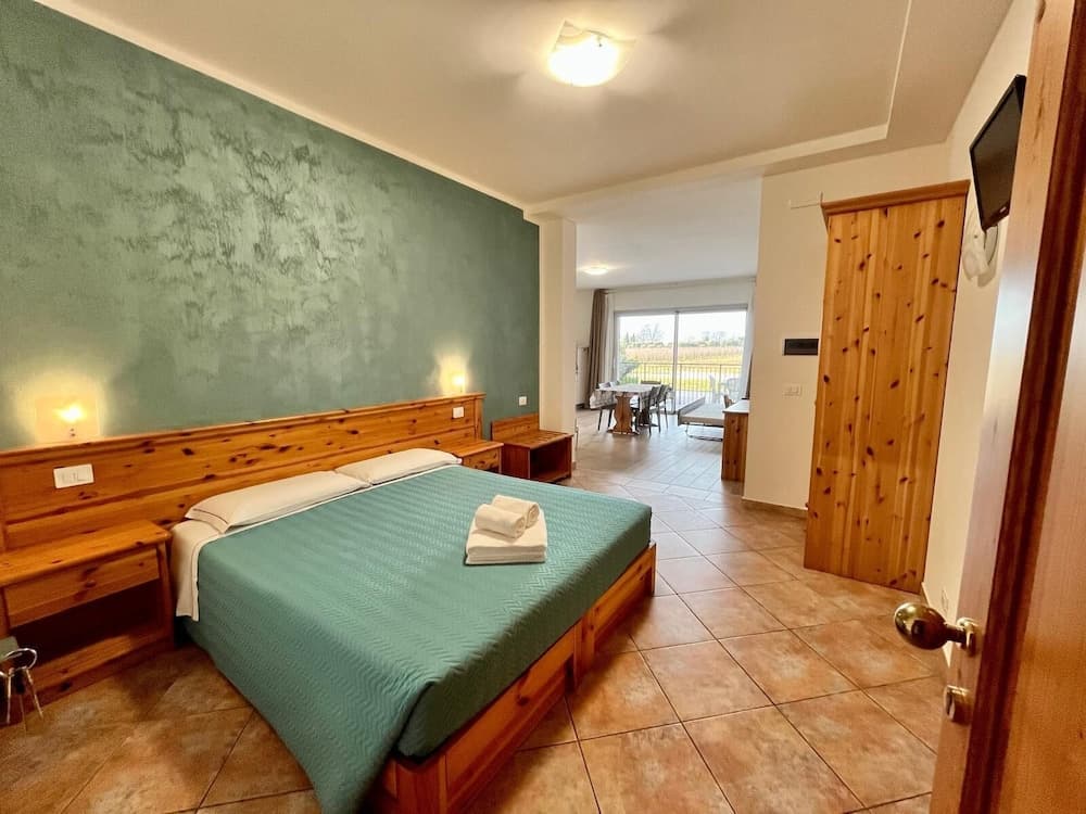 A cozy hotel suite features a neatly made bed with green linens, wooden furnishings, and a dining area leading to a balcony, ideal for travelers seeking comfort and relaxation.