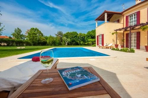 A serene villa scene features a tranquil poolside area with lounge chairs, a glass of wine, and a travel book on a wooden table, set against lush greenery and a charming house, ideal for a relaxing vacation getaway.
