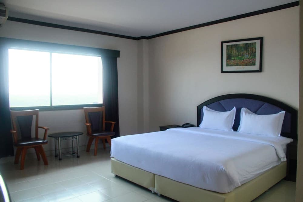A cozy hotel room features a neatly made bed, two chairs by a bright window, and a framed forest painting, ideal for travelers seeking comfort and simplicity.