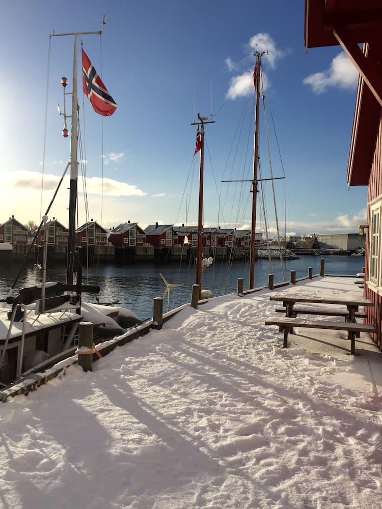 A serene winter scene at a snow-covered dock by the water, featuring traditional red houses, Norwegian flags, and sailboats, ideal for travelers seeking a picturesque Nordic coastal experience.