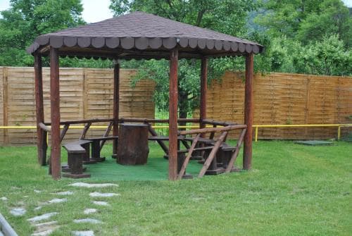 A serene outdoor picnic area features a wooden gazebo with rustic benches on a grassy lawn, ideal for family gatherings or nature retreats.
