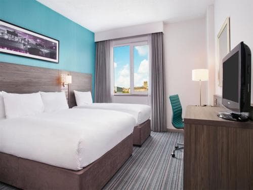 A cozy hotel room with a neatly made bed, vibrant blue wall, and a desk area by the window offers a comfortable stay for travelers seeking relaxation and convenience.