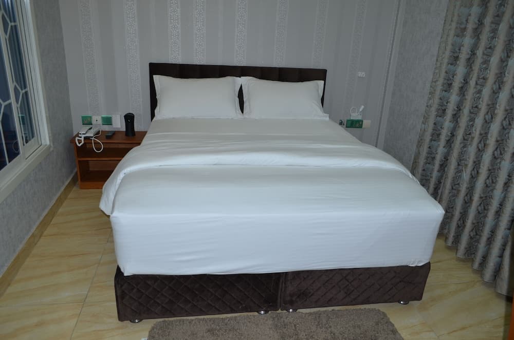 A cozy hotel room features a neatly made bed with white linens, patterned wallpaper, and a soft rug, ideal for travelers seeking comfort and relaxation.